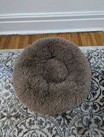Fluffy brown circular plush pet bed on patterned rug, showing its nest-like padded design from front perspective.
