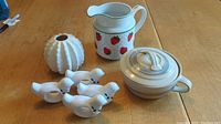 Photo of all ceramic items arranged on wooden table - bud vase, strawberry pitcher, beige bowl with lid, and four bird-shaped napkin holders.