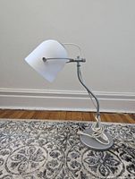 Front view of the Ikea desk lamp showing the white cone-shaped shade angled forward, metal curved arm, round base, and coiled white cord on patterned carpet.