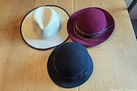 Photo showing three hats on a table: black wool bowler, burgundy wool fedora, and cream straw hat with black trim.