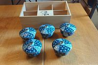 Set of five blue and white Japanese ceramic bowls with lids arranged in front of a wooden storage box with compartments on a wooden table.