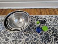 Three nested stainless steel mixing bowls with five assorted reusable plastic mason jar lids on a patterned rug.