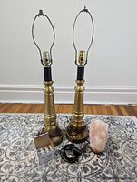 Photo of two brass lamps with harps, a Himalayan salt lamp, electrical cords, and a box of four Harage lightbulbs on a patterned rug near a baseboard.