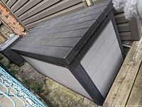 Outdoor storage box showing black lid and gray side panels, placed next to house and chain link fence.