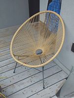 Photo shows one round patio chair with woven beige rattan seat and backrest, standing on a wooden deck floor near a grey wall.