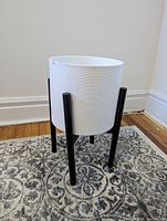 Front view of the white cylindrical plant pot set on a black three-legged stand placed on a patterned rug against a wall.