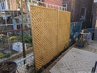 Wide angle photo showing two connected wooden lattice privacy screen panels installed along a fenced outdoor area with a patio rug in front.