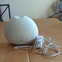 Closed view of round white Saje Aroma Breeze diffuser with power cord and adapter on wooden surface.