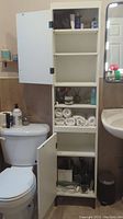 Shelving unit with both doors open showing multiple shelves, some with bathroom items and folded towels, placed in bathroom near toilet and sink.