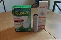 Box of Polident Retainer Cleanser tablets, unopened Bio-Oil bottle, and Saje Eater's Digest remedy stick on a wooden table.