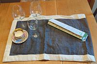 Three wine glasses (two red wine glasses and one smaller glass), floral teacup with matching saucer, Abeego beeswax food wrap in packaging, and dark blue handwoven table runner with white border on wooden table.