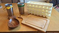 Two stainless steel travel mugs with yellow and green bands, wooden pineapple-shaped bowl, wooden rectangular cutting board, and four white plastic ice cube trays arranged on wooden table.