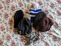 Five winter hats and one pair of gloves laid out on floral fabric background. Hats include knit and felt styles, one with fur pompom, and gloves have decorative bow detail.