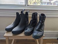 Two pairs of women's boots on wooden stool by window. One pair black leather ankle boots, creased and scuffed. Other pair black rubber rain boots.