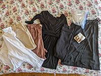 Collection of women's clothing in neutral tones spread on floral fabric background showing pants, tops, black dress, and additional black clothing items with a small package.