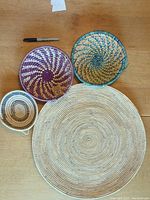 Four woven grass baskets arranged on a wooden surface with a marker for size reference. Shows the purple basket with some fading and detail of colors and patterns.