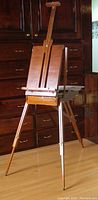 Photo of fully assembled wooden folding easel standing on floor with tripod legs extended and canvas clamp at the top.