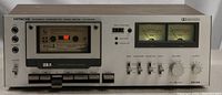 Front angle of Hitachi stereo cassette tape deck powered on showing cassette compartment with tape inside, analog VU meters lit up, and control panel.