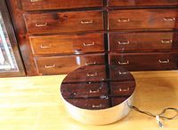 Top round metal display stand with black reflective top and silver sides, showing minor surface wear and electrical cord extending from base.