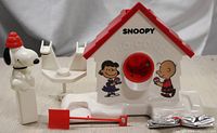 Front view of the Snoopy Sno-Cone machine with Snoopy figure, clamp accessory, and red shovel visible on a surface.