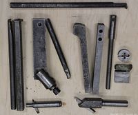 Top view of approximately 12 metal lathe accessories including boring bars, tool rests, rods, die, and holders scattered on plywood surface.