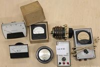 Vintage electronic meters with analog dials and needles, a boxed meter, rotary switch, and a signal tracer device shown together on a light wood surface.