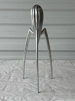 Front view showing the metal citrus squeezer on its three long angular legs, with the ridged oval cone used for pressing citrus at the top.