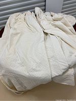 Photo showing heavy cream cotton drapery panels folded and draped. Panels have subtle floral/damask texture, showing folds and light discoloration spots.