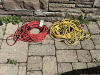 Two coiled electrical extension cords, one red and one yellow, lying on a stone pavement against a stone wall background.