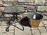 Full view of square glass top patio table with black metal legs alongside modern abstract gold metal planter with gray interior.