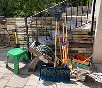 Front view of full gardening lot showing green garden hose on brown rolling hose reel, two shovels with wooden handles, two rakes, broom, green plastic garden stool, box of firewood, and small tools in containers.