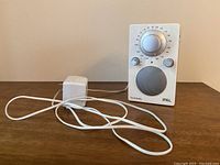 Front view of white Tivoli Audio iPal portable radio alongside its white power adapter with attached cord, placed on a wooden surface.