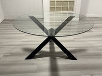 Front view of modern round clear glass table with black metal X tripod legs on light wood floor.