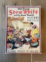 Top view of original puzzle box showing artwork of Snow White and the Seven Dwarfs gathered around a fireplace, with clear visible printing of the puzzle title and publisher on the box top.