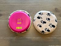 Two circular SnuggleSafe heat pads for pets, one showing pink underside with warning labels, the other covered with beige fabric featuring black paw prints, placed side by side on wooden surface.