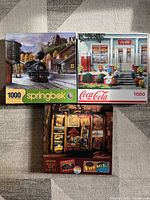 Three puzzle boxes laid flat on carpet floor. Top left: 1000 piece train station puzzle by Springbok. Top right: 1000 piece Coca-Cola puzzle. Bottom center: 500 piece vintage signs puzzle.