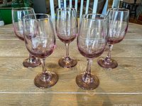 Front and side view of 5 pink tinted wine glasses arranged in a semi-circle on wooden surface.