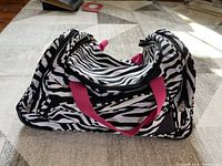 Zebra print soft sided duffle bag with pink handles showing front with zipper pocket and side handles.