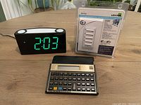 Photo shows digital alarm clock displaying 2:03 with large green numbers, HP 12C calculator with black body and keypad, and unopened Leviton 30-minute digital timer in packaging