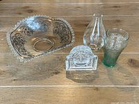 Four glass items on a wooden surface: ornate fancy bowl, decorative glass napkin holder, vintage green Coke glass tumbler, and clear crystal vase.