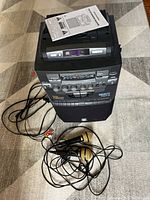 Photo of the karaoke machine with two wired microphones and cables placed in front of it on a carpeted floor. The machine is black with various buttons and dials, including a cassette tape deck.