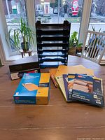 Seven black stacking organizers displayed on a wooden table in front of window, with two binders and various mailing envelopes and label packages placed in front.