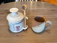 Both pottery items together shown on a table: the olive pot with attached wooden scoop and the mug.