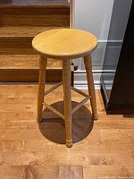 Front angle view of the solid oak stool placed on wooden floor showing round top and four legs with supporting bars.