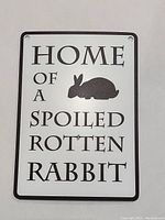 Photo of a white rectangular metal hanging sign with black border, black text, and rabbit silhouette, showing holes at top for hanging.