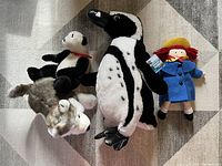 Plush animals and dolls arranged on gray patterned surface including large penguin, smaller penguins, Madeline doll, wolf and panda plushes.