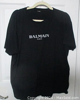 Front view of black Balmain Paris t-shirt showing white logo in center chest area, hung on hanger against white door.