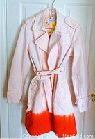 Front view of the white denim trenchcoat showing the ombre orange hem, belt tied at waist, collar and double-breasted button layout.