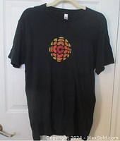 Full frontal photo of black T-shirt with CBC vintage logo hung on a door.