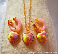 Photo showing gold-tone necklace with tie-dye heart charm with eagle emblem and two matching pink huggie earrings with dangling heart charms.